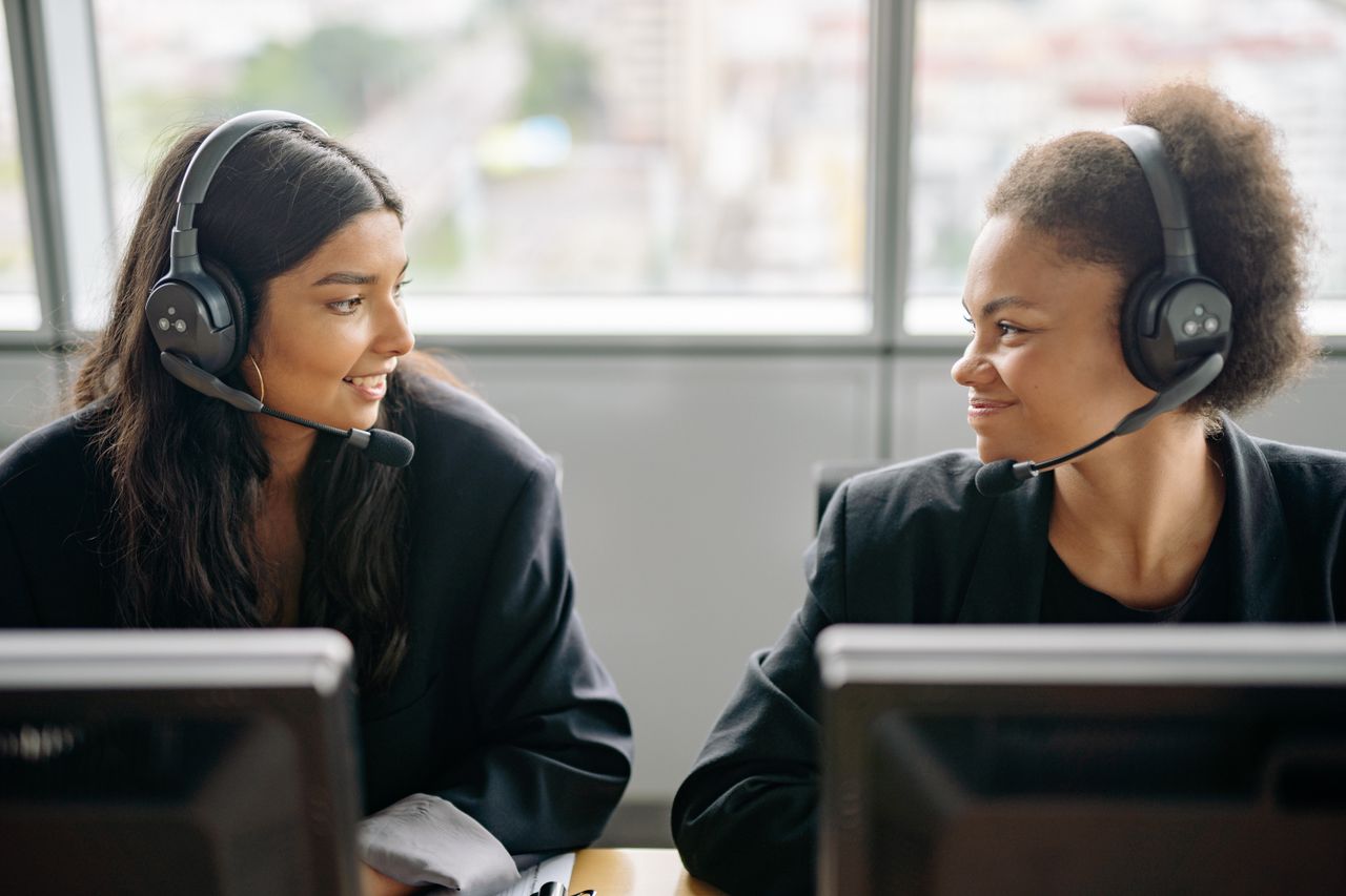 who-we-are Two smiling female call center agents with headsets engaging in teamwork.